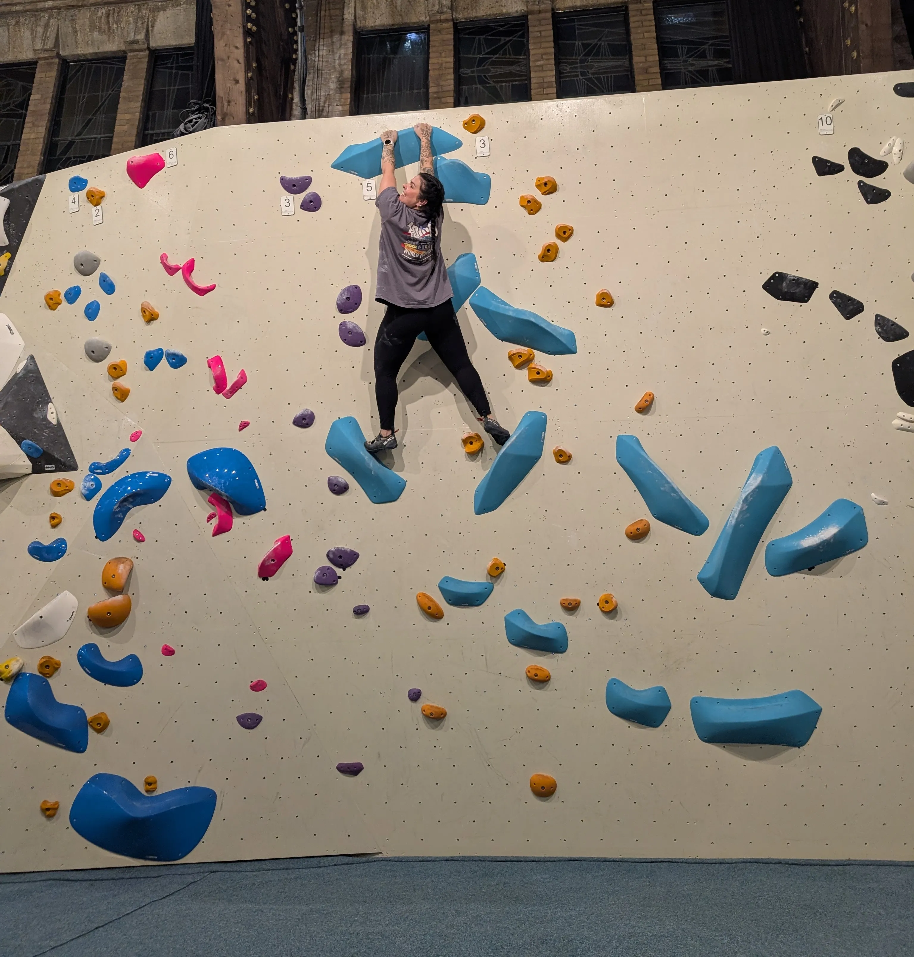The image shows an indoor climbing wall with colored boulders. I stand on top of two big blue holds while holding with both hands a big blue holder that is the end of that route. The routes have different numbers, and the one I am finishing is a 5.