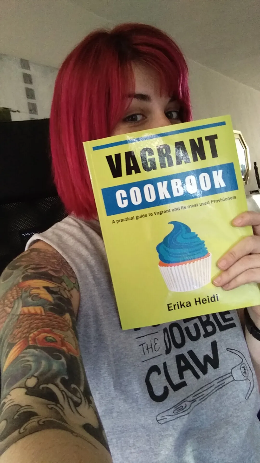 Picture showing me in 2015 with a vibrant pink/red short hair holding a book in front of me, covering part of my face. The book has a bright yellow cover and has the title "Vagrant Cookbook" with black and blue thick letters, and a cupcake illustration.
